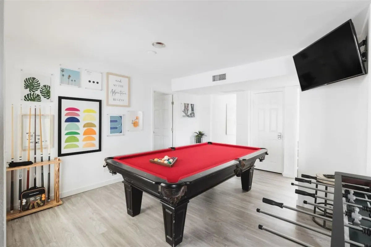 A casual game room featuring a pool table with red felt, colorful wall art, a flat-screen TV, and billiard cues. The space is designed for fun hangouts, hosting game nights, and relaxed socializing with friends.