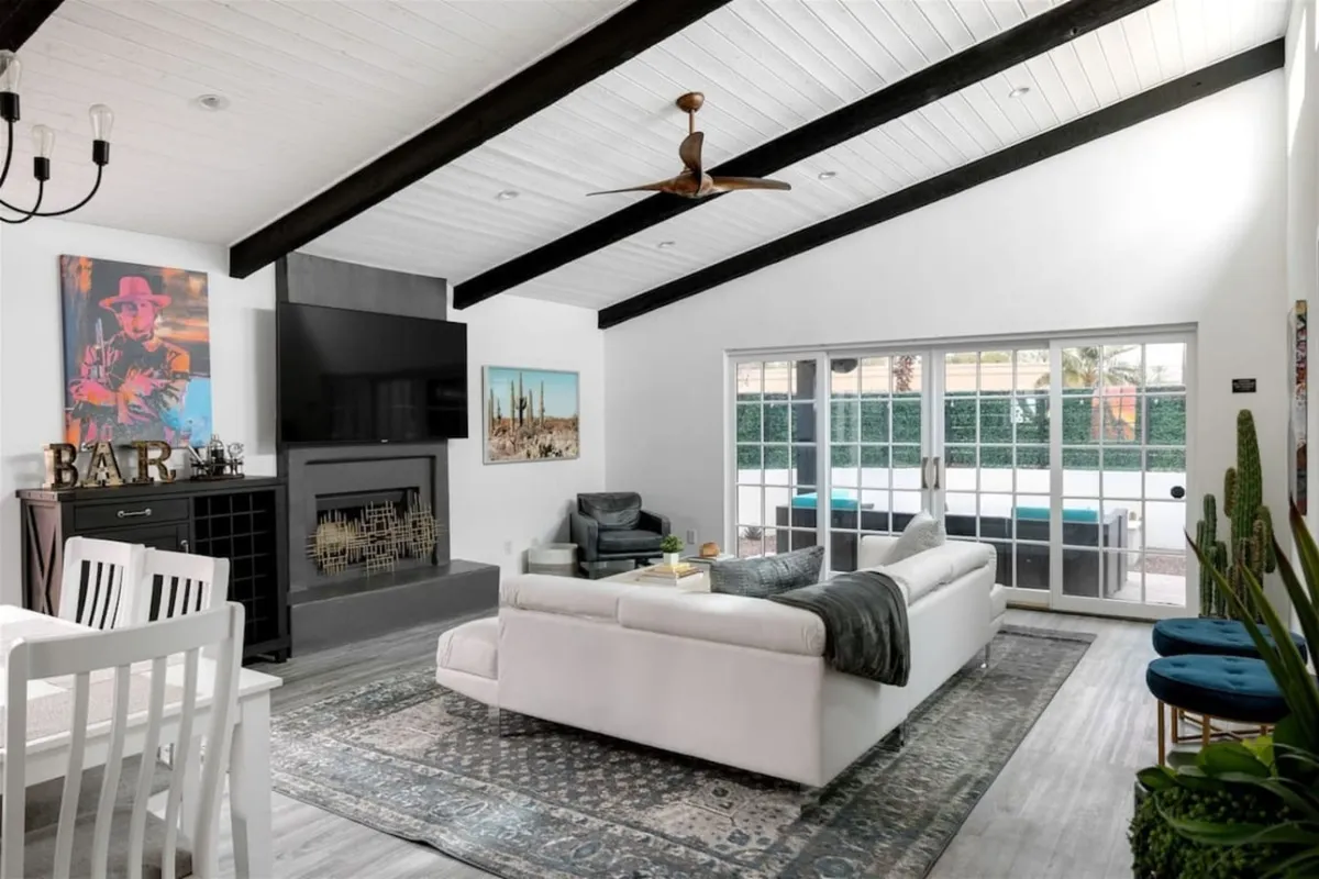 A spacious, light-filled living room with a large white sectional sofa facing a wall-mounted TV above a sleek fireplace. Nearby, a dining table with chairs provides additional gathering space, and colorful artwork adds vibrancy. Natural light streams in through large windows, creating an inviting setting for relaxation or entertainment.