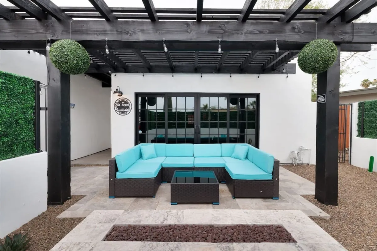 An inviting outdoor seating area under a pergola, surrounded by lush greenery. The cozy space features a coffee table and comfortable chairs, with string lights overhead creating a festive atmosphere, perfect for socializing or relaxing. The area offers lovely sunset views and is ideal for casual brunches or evening gatherings.