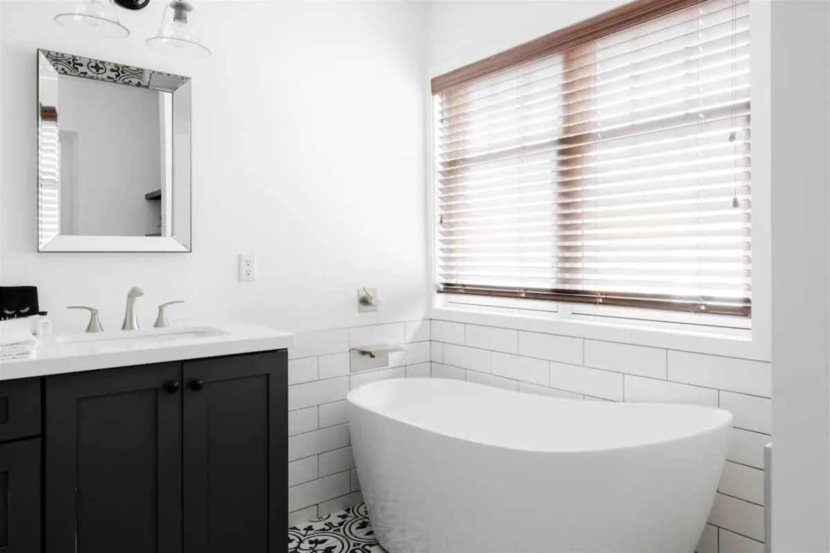 A spa-inspired bathroom with a sleek freestanding bathtub, modern vanity and sink, a large mirror, and a sunlit window with blinds. The space feels airy and luxurious, perfect for self-care routines and rejuvenation.