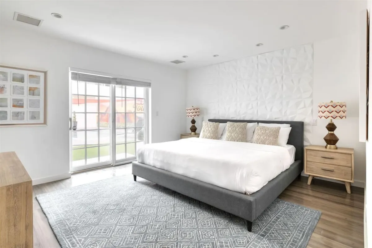 A serene, light-filled bedroom featuring a plush bed set against a decorative wall, bedside tables, sliding glass doors that open to the outdoors, and a cozy area rug. The space feels tranquil and welcoming, perfect for unwinding after a busy day.