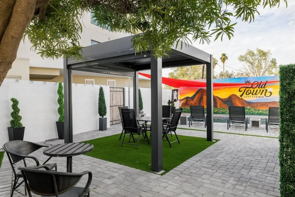 A lively outdoor seating area shaded by a pergola, adorned with a colorful mural and surrounded by lush greenery. The space is arranged for both communal dining and social gatherings, with art as a backdrop to relaxation and conversation.