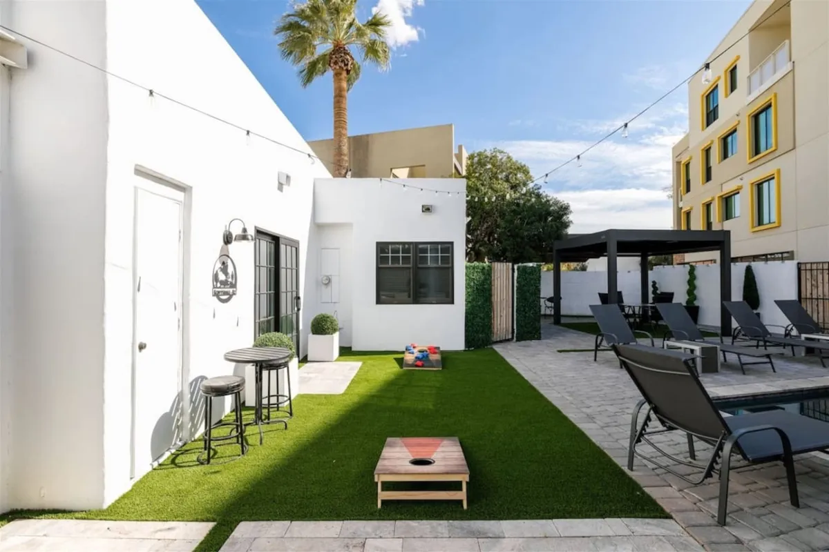 A lawn area adjacent to the pool with outdoor seating, decorative lighting, and a palm tree, set for casual fun. People are sunbathing, playing cornhole, socializing, and enjoying outdoor dining as the sun sets, evoking a festive, communal vibe.