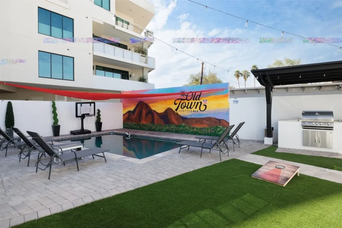 A vibrant backyard scene featuring a sparkling swimming pool, a colorful mural wall, outdoor seating, a dedicated grill area, and a lawn set up for games. The area is bustling with activity, inviting families to swim, barbecue, sunbathe, and play together in a lively, communal setting.