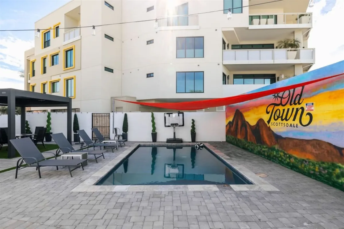 A vibrant outdoor pool area featuring a colorful mural as a backdrop, with stylish lounge chairs, a cabana structure, and groups of friends relaxing by the water. Soft sunset lighting enhances the festive mood, highlighting the inviting space for social gatherings and evening events.