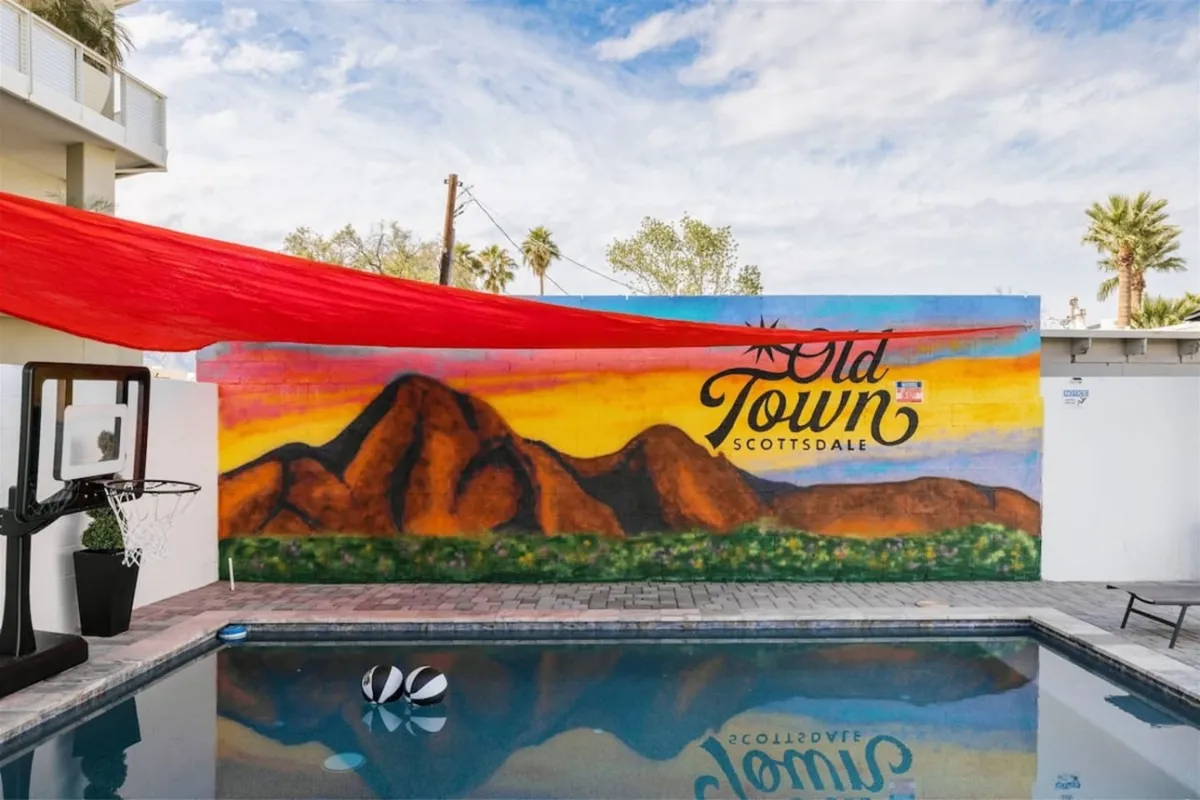 A vibrant backyard scene showcasing a sparkling swimming pool with a basketball hoop, a playful colorful mural as a backdrop, and a shaded red fabric canopy. Friends are swimming, sunbathing, and socializing poolside, while others enjoy the mural and engage in lighthearted activities.